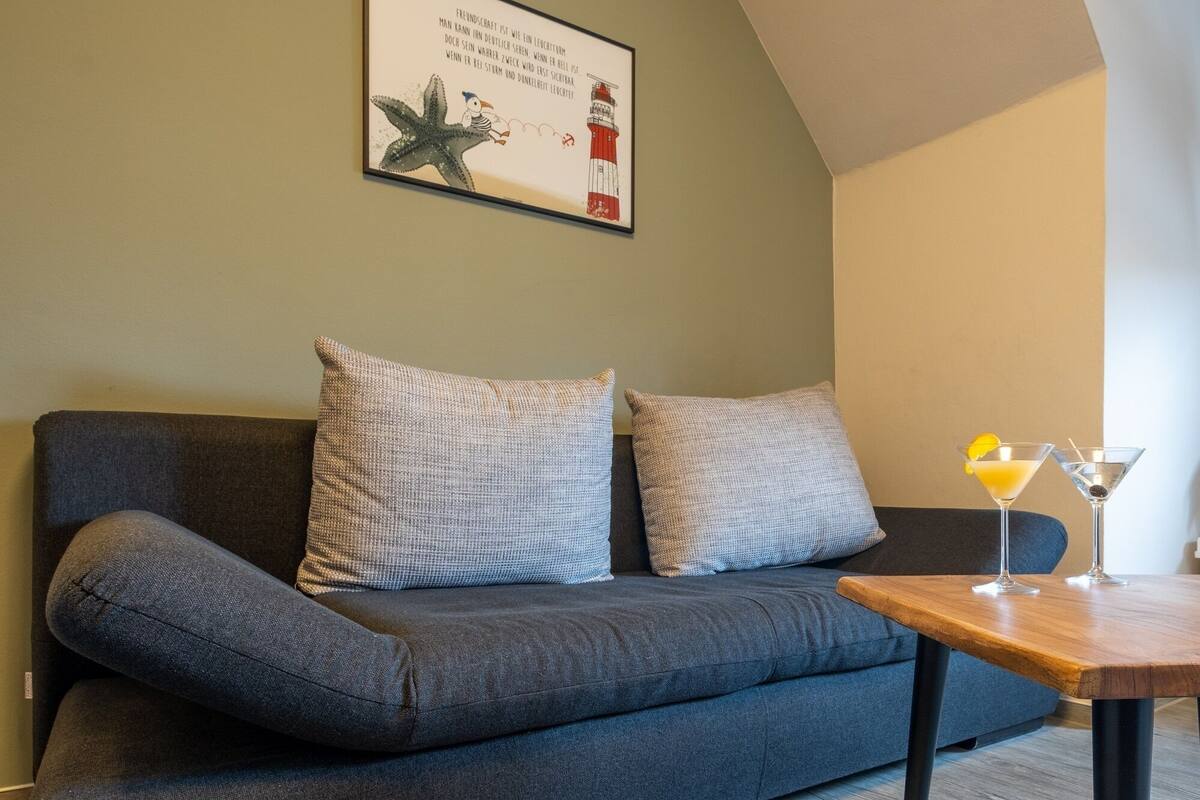 Un salon moderne avec un canapé gris foncé, deux coussins à motifs, une œuvre d’art encadrée au-dessus du canapé et une table basse en bois sur laquelle se trouve un verre à martini.