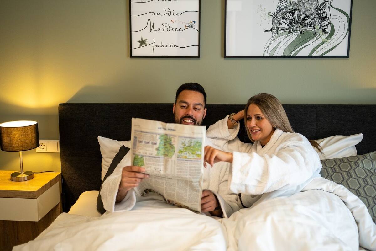 Un couple est assis dans une chambre d’hôtel, lisant un journal. Une lampe est posée sur une table de chevet et une œuvre d’art orne le mur.
