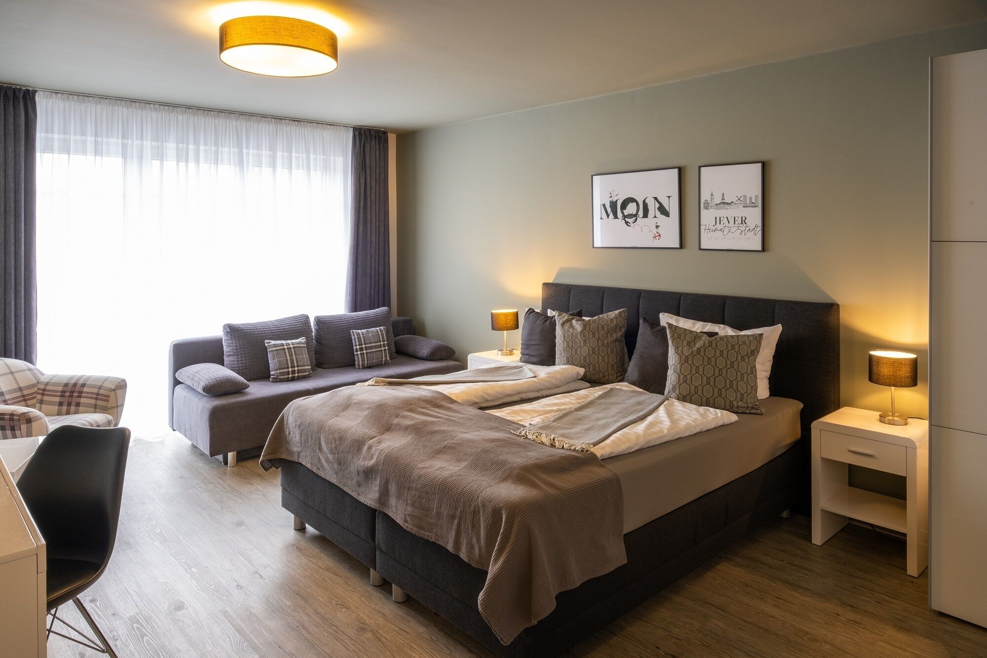 A modern hotel room with a large bed, bedside tables, a sofa, and a chair. There are two framed pictures on the wall and a window with curtains.