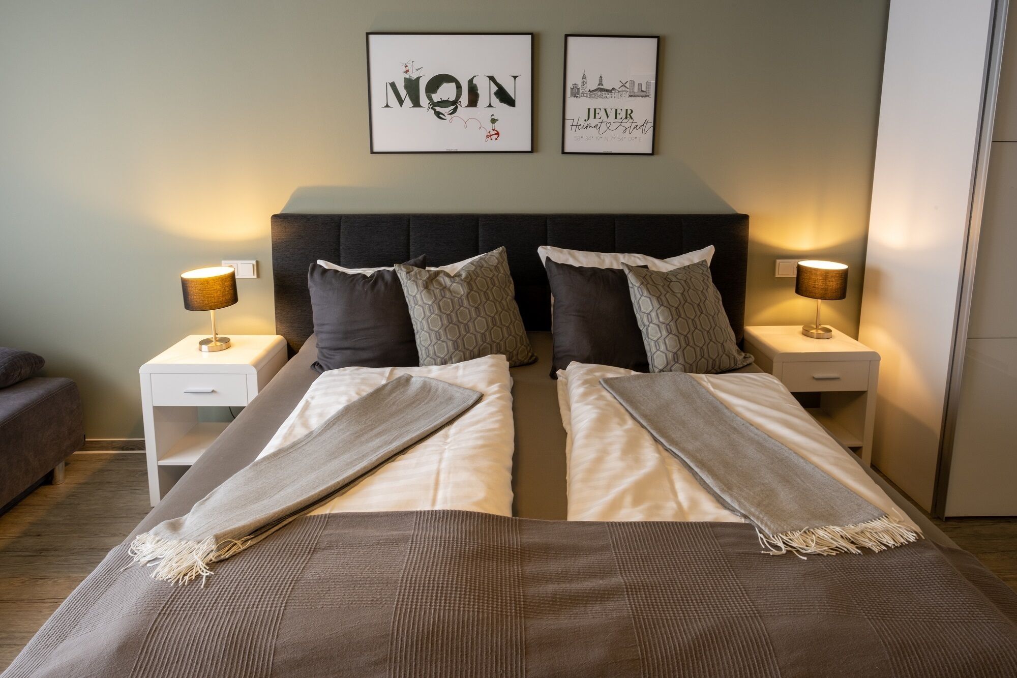 Uma cama de casal com edredom cinza e branco, duas mesas de cabeceira com luminárias, e duas obras de arte emolduradas na parede.