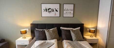 Una cama doble con edredón gris y blanco, dos mesitas de noche con lámparas y dos cuadros enmarcados en la pared.