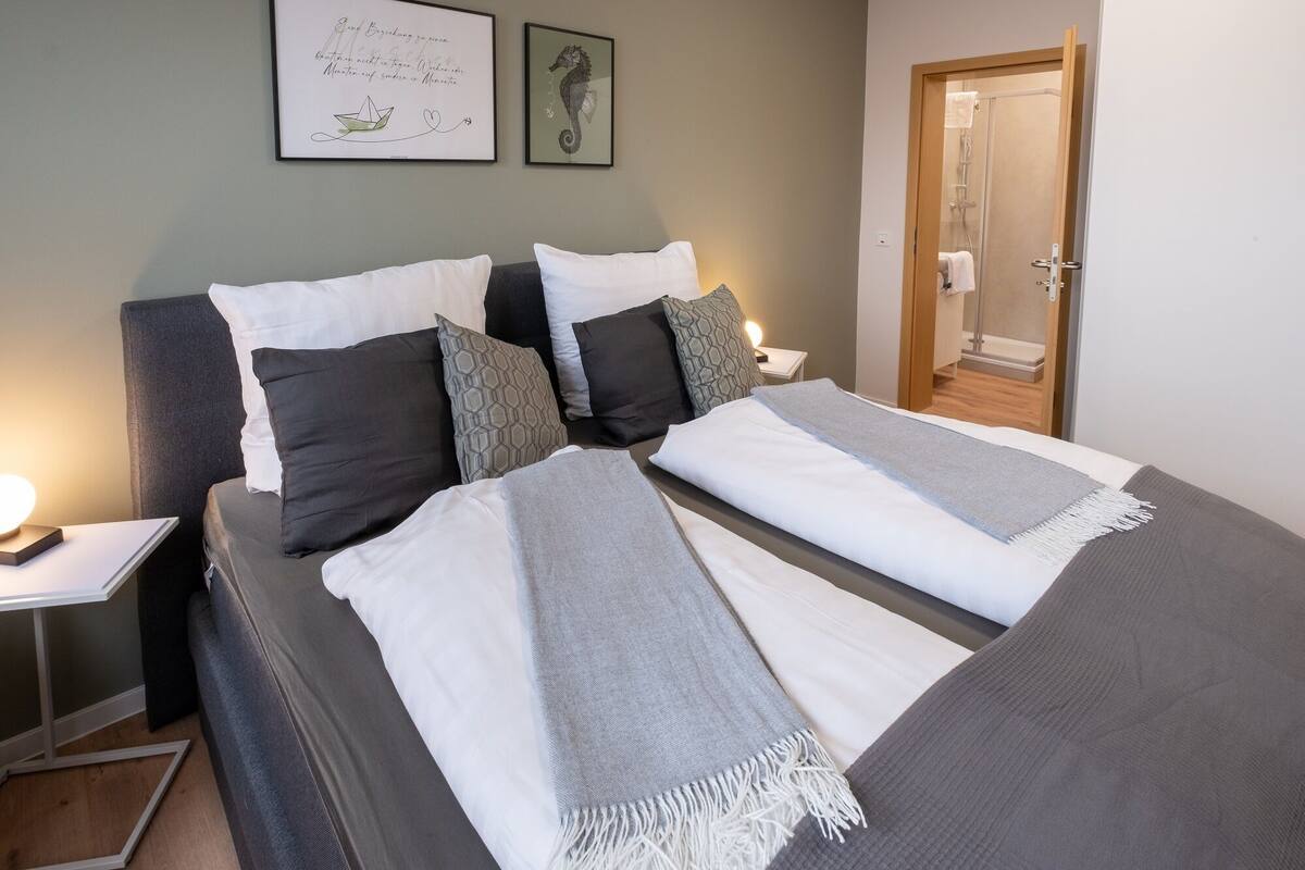 Un lit bien fait, recouvert d’une couverture grise et blanche, avec un oreiller gris et un oreiller blanc. Un tableau encadré est accroché au mur, et une porte se trouve sur le côté droit.