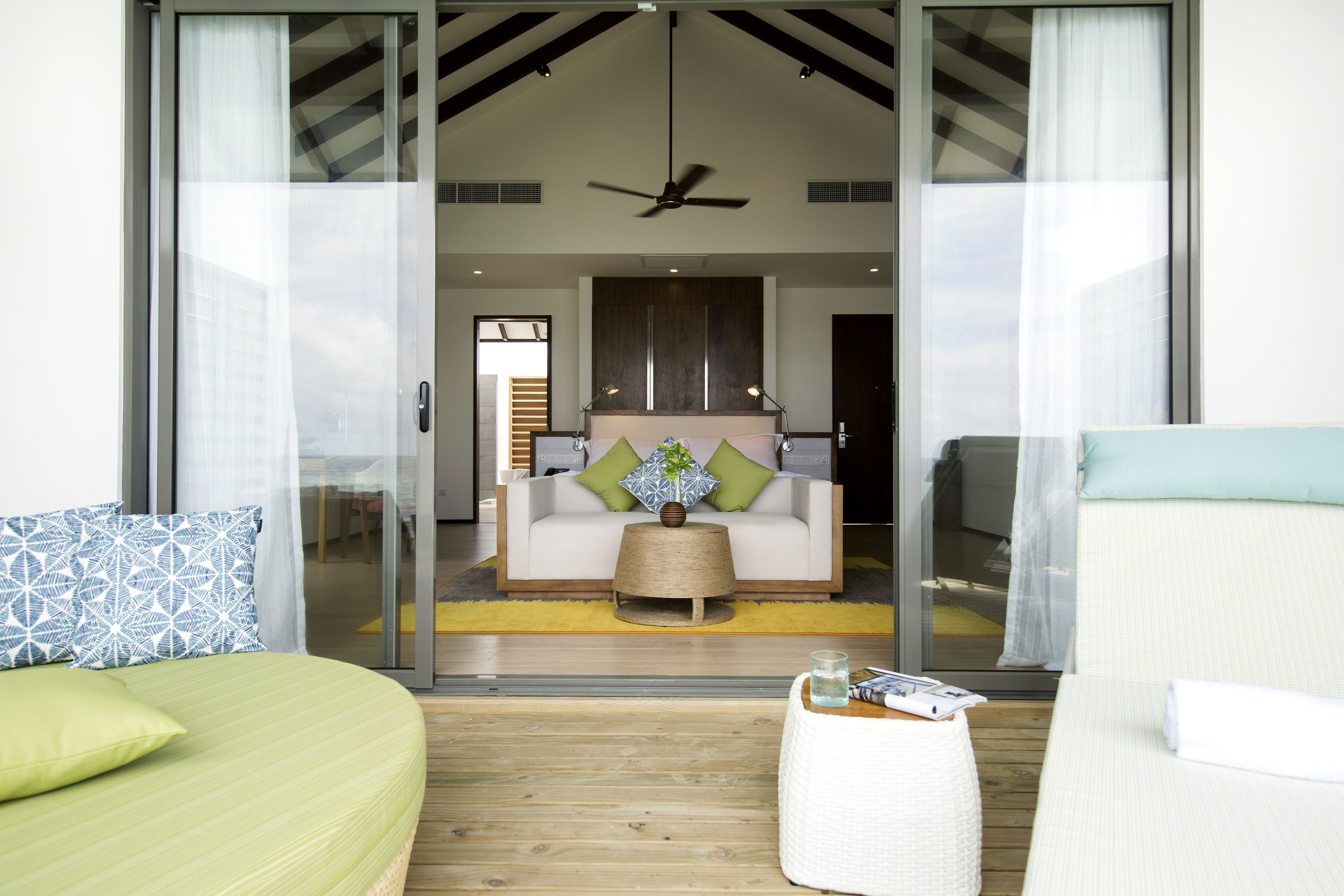 A modern hotel room with a large sliding glass door leading to a balcony, a sofa, a small side table, and a bed with green bedding.