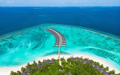 An aerial view of a resort with overwater bungalows connected by a walkway, surrounded by turquoise waters and a white sandy beach.