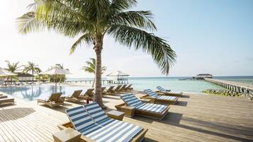 A wooden deck with lounge chairs and a pool, palm trees, and a pier extending into the sea.