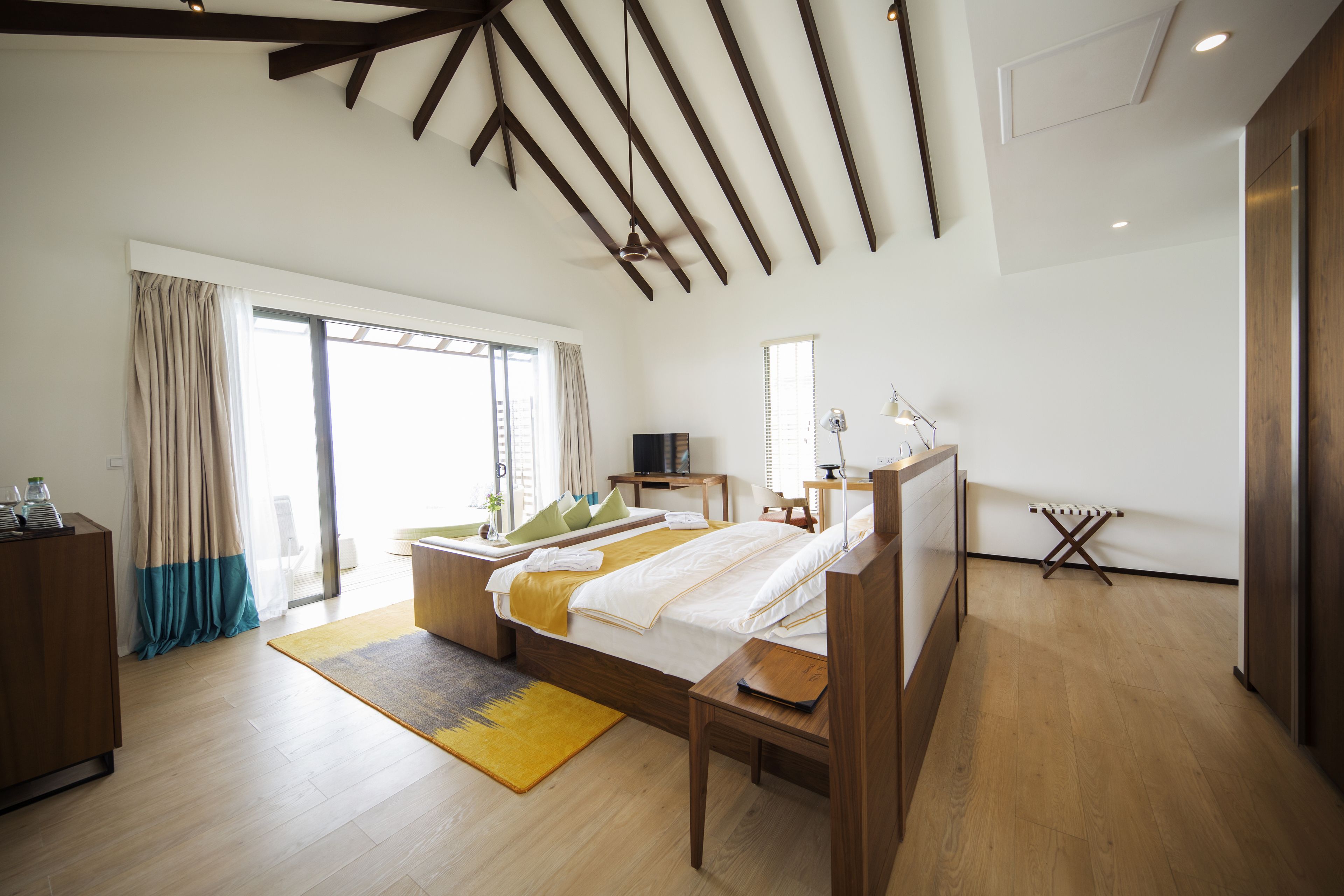 A modern bedroom with a large bed, wooden beams on the ceiling, a desk, and a window with curtains.