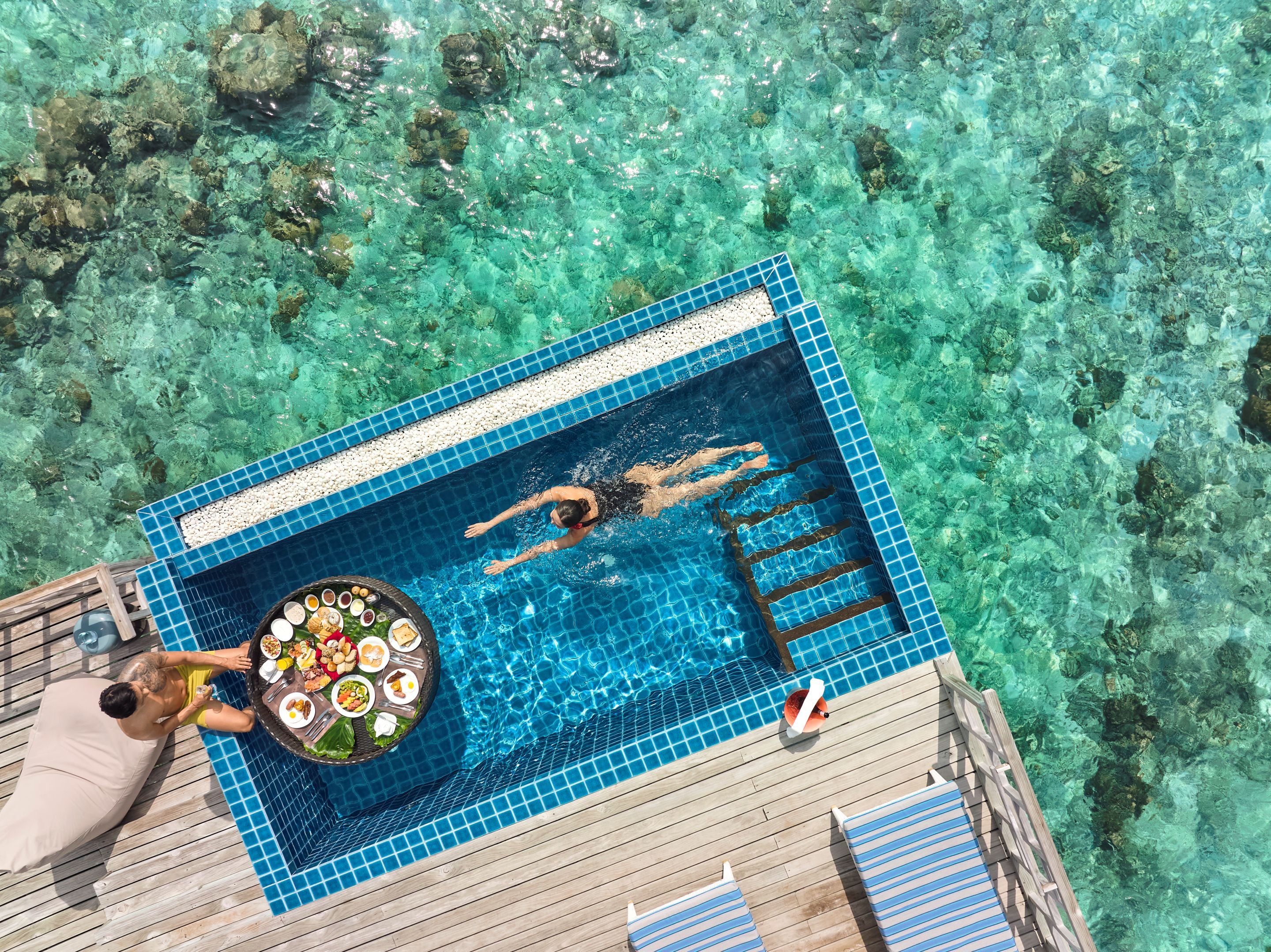 Vista aérea de una piscina con agua cristalina, una mesa de comedor con comida y sillas de descanso.