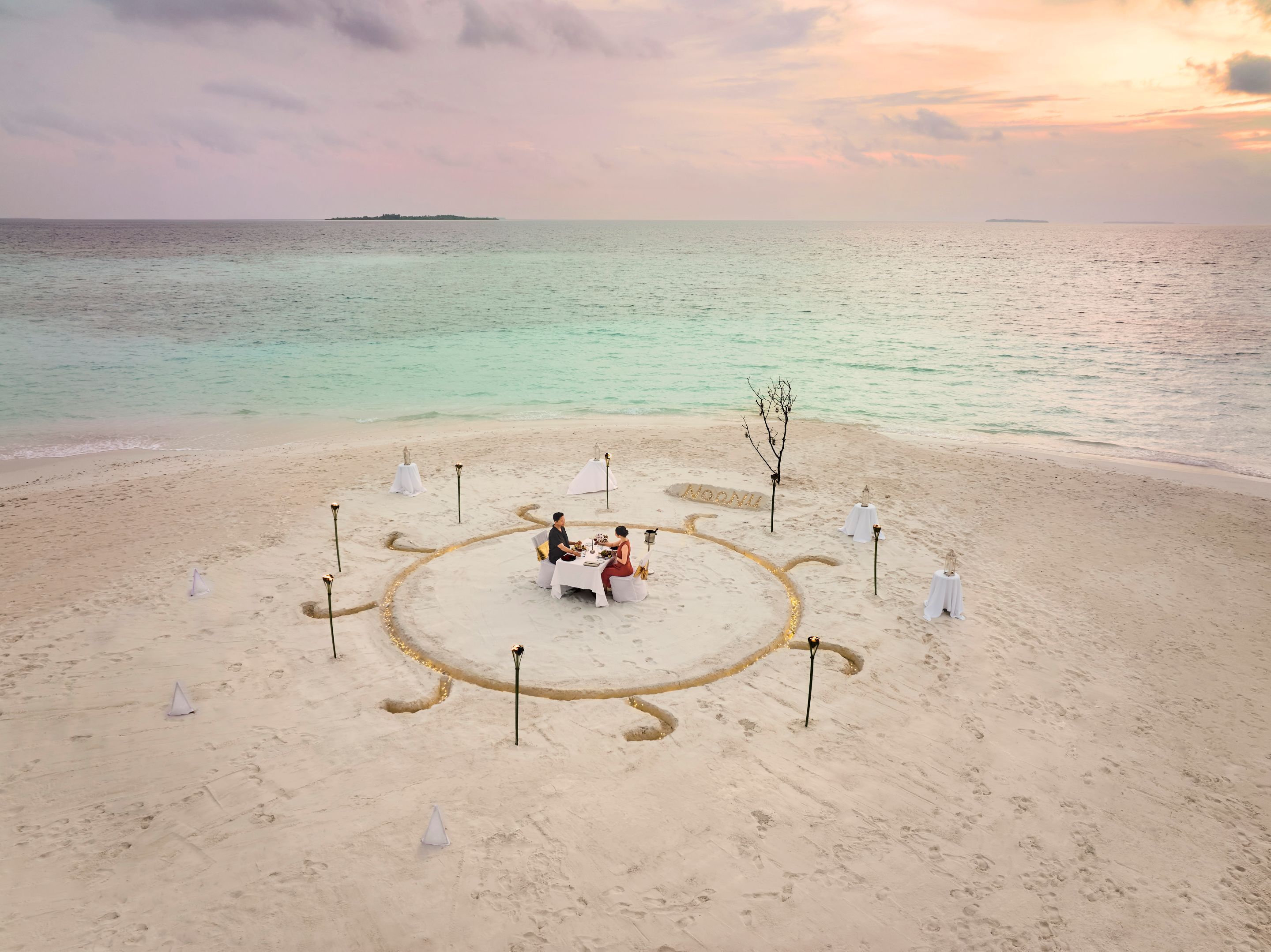 A beach setting with a circular arrangement of tables and chairs, a tree, and a small island in the distance.