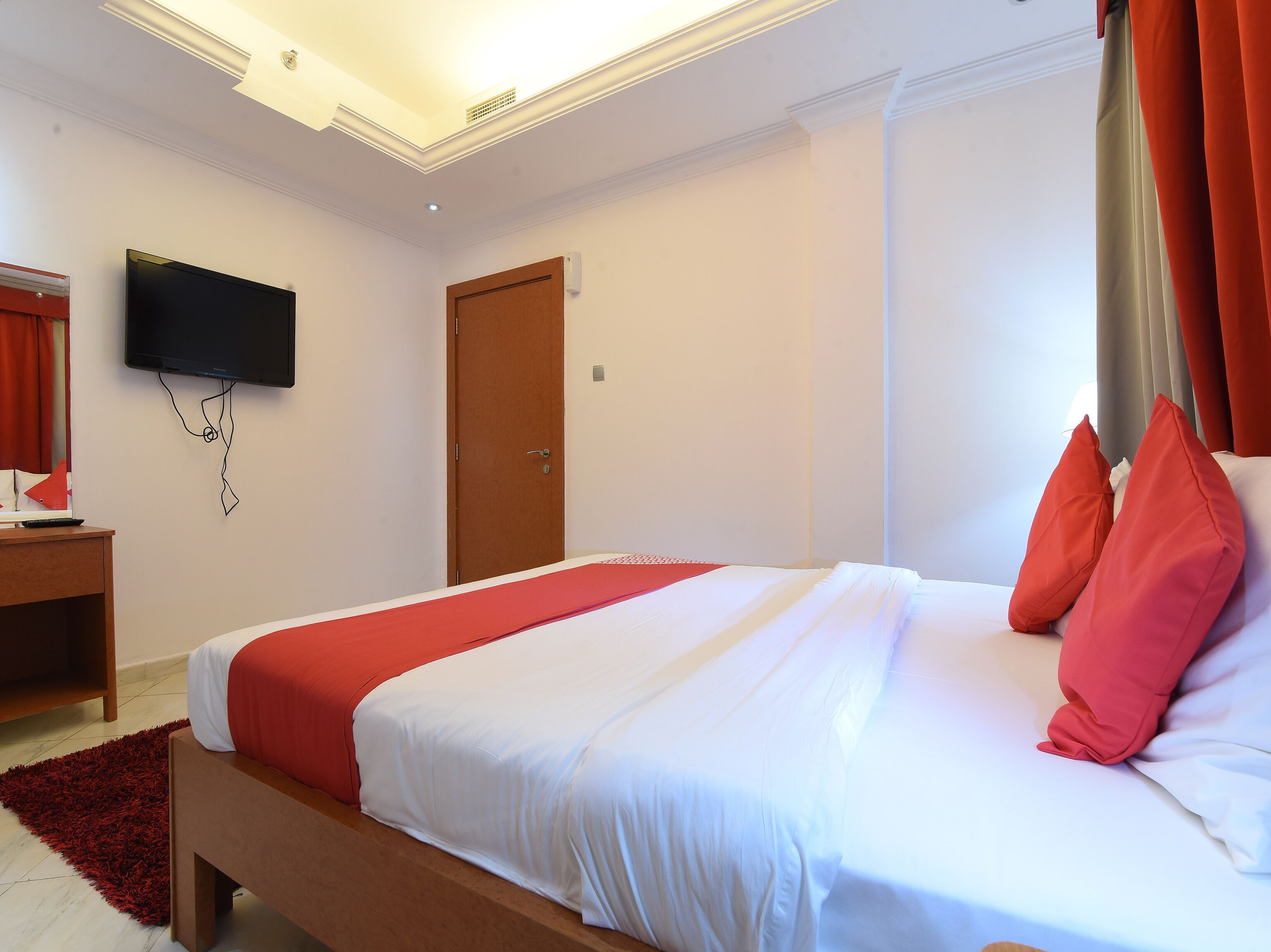 Una habitación de hotel con una cama, un televisor montado en la pared, una mesita de noche de madera y una alfombra roja.