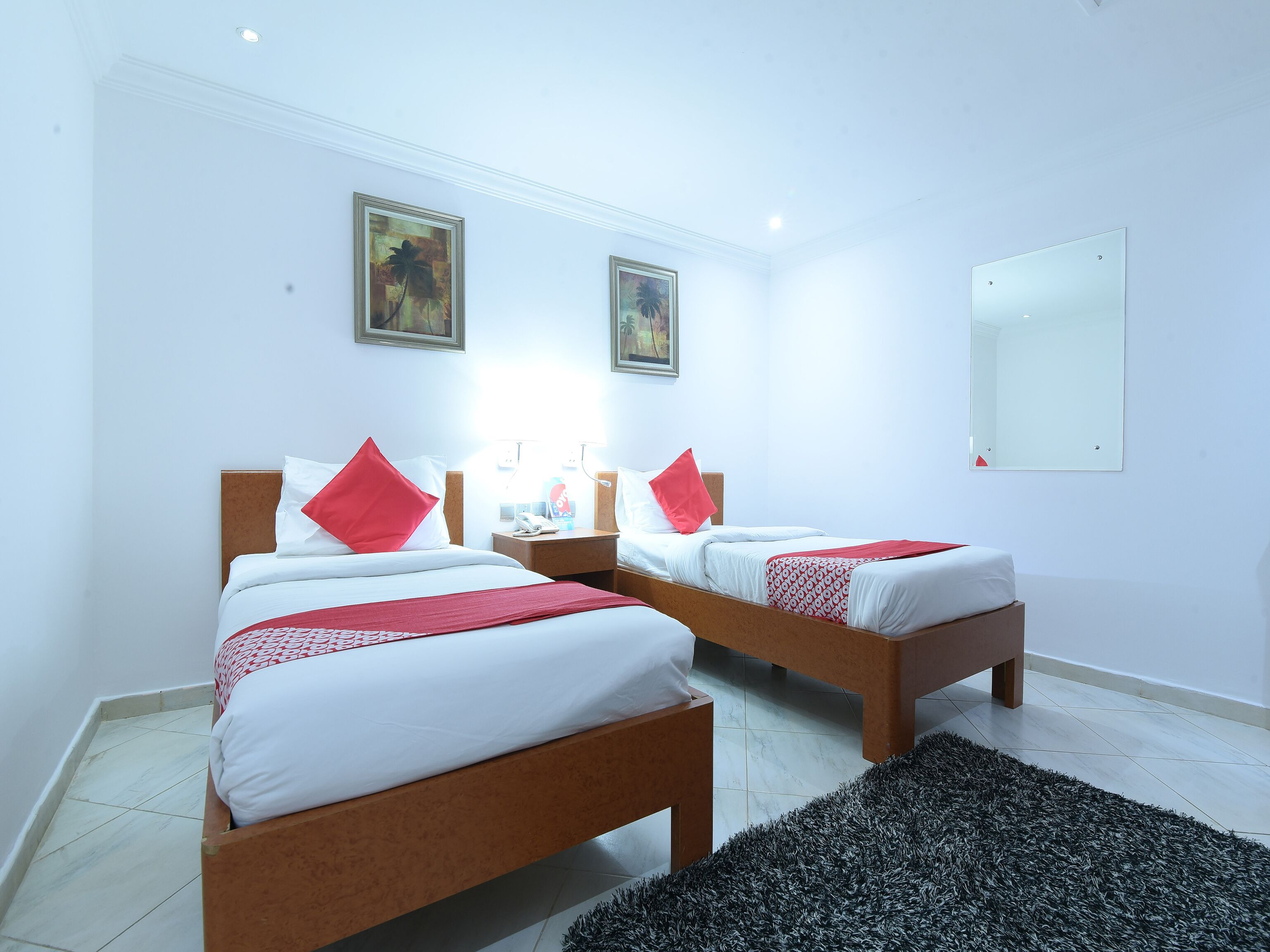 Habitación de hotel con dos camas, ropa de cama blanca, almohadas rojas, alfombra blanco y negro y dos cuadros en la pared.
