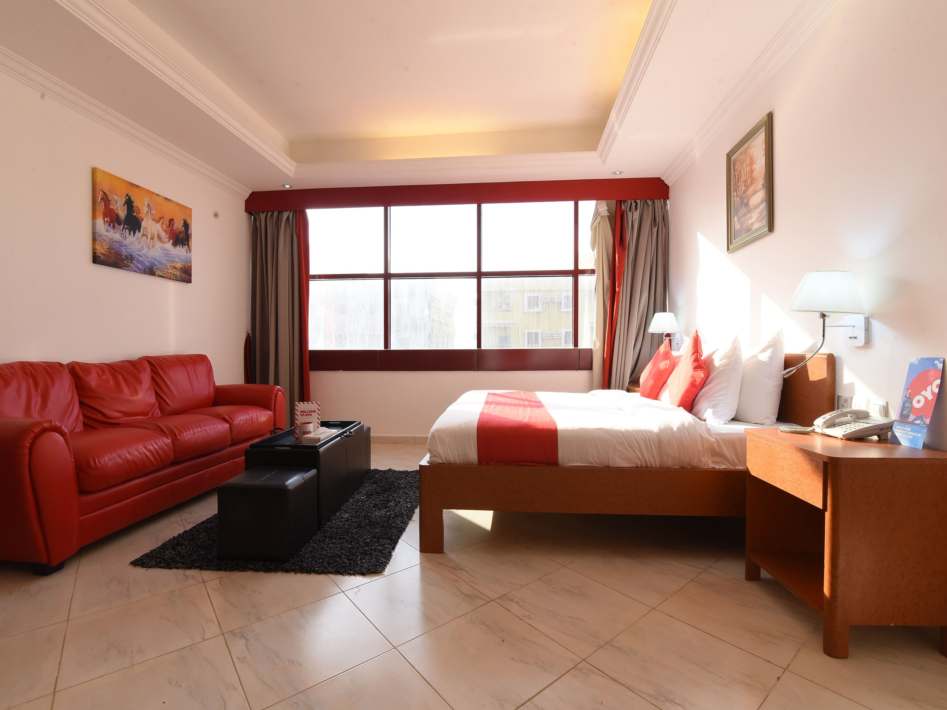 Habitación de hotel con una cama, un sofá rojo, una mesita, una mesilla de noche, una lámpara y un cuadro en la pared.