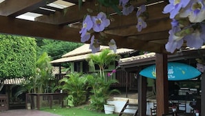 An outdoor pool area with a wooden deck, surrounded by lush greenery and a covered seating area with a bar. - Pousada Aloha Beach House (Imbituba)