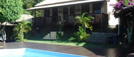 A poolside area with a wooden deck, a pool, and a house with a tiled roof and a porch.