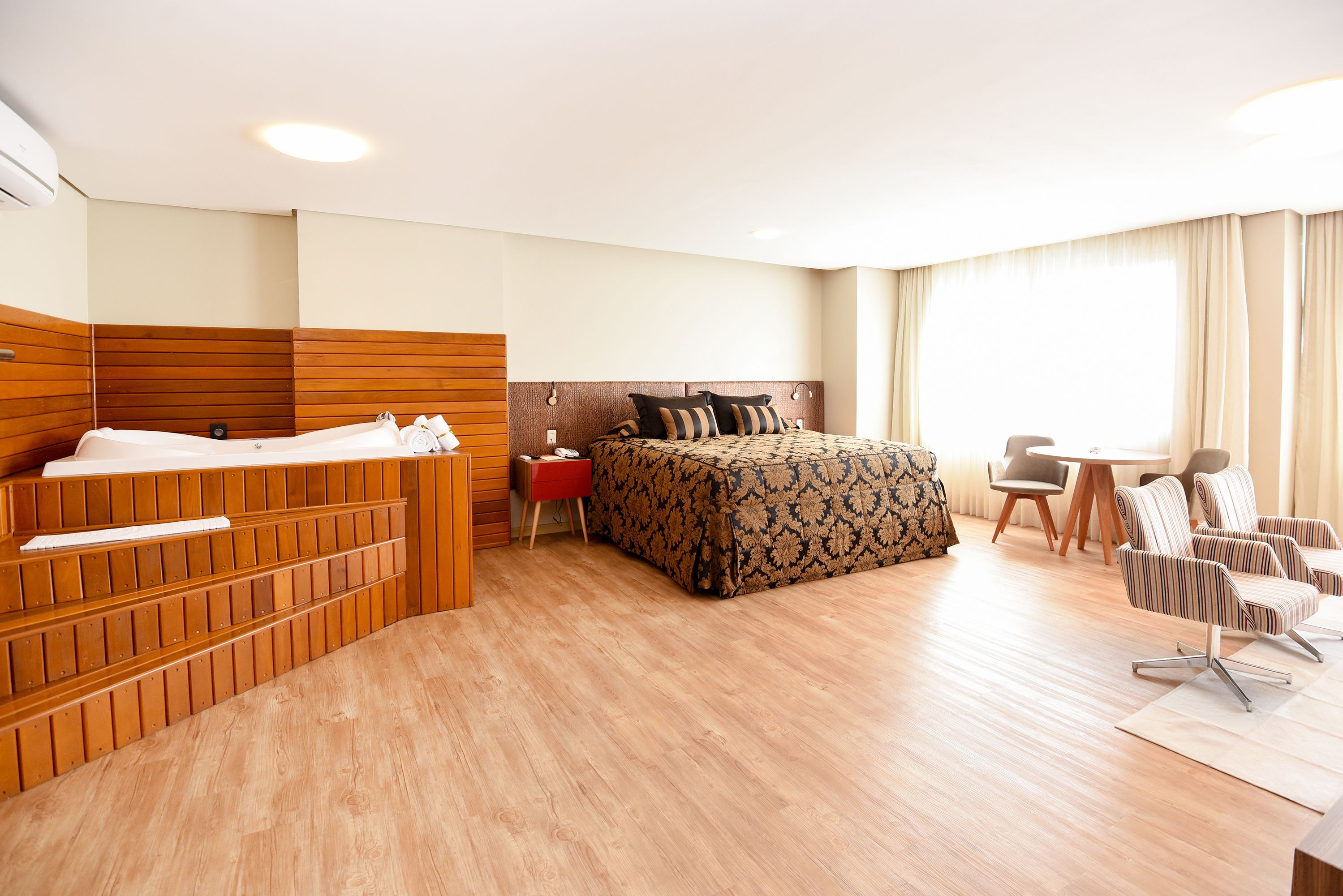 Une chambre d’hôtel avec une tête de lit en bois, un lit avec une literie à rayures, un petit bureau, une chaise et une table ronde.