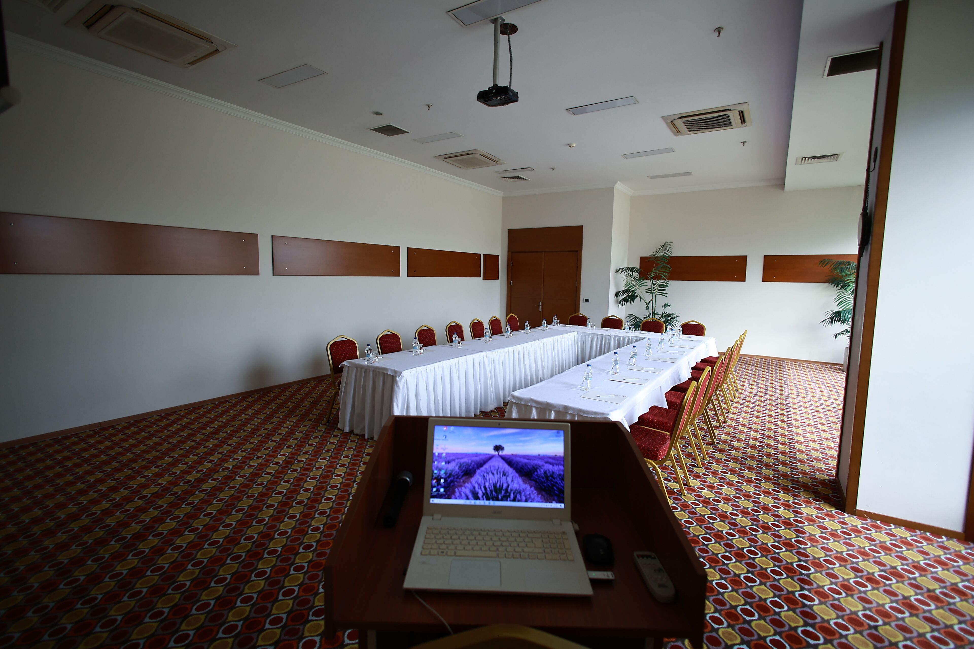 A conference room with a long table, red chairs, a laptop, and a patterned carpet.