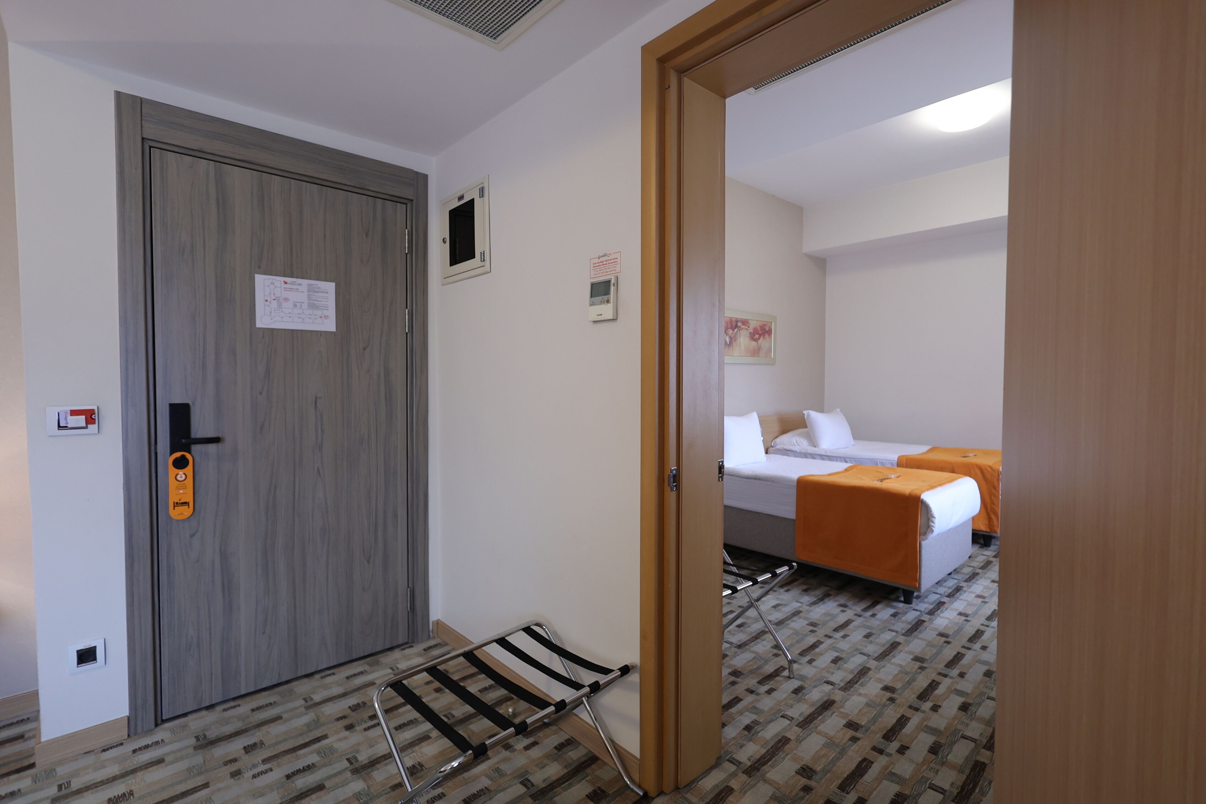 A hotel room with a wooden door, a bed with an orange bedspread, a small table, and a wall-mounted heater.