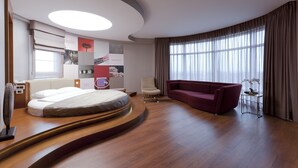Quarto de hotel moderno com uma cama curva, um sofá vermelho, uma mesinha com um vaso e uma janela grande com cortinas.