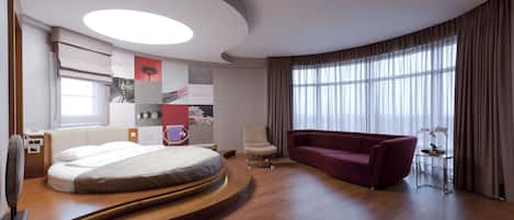 Una habitación de hotel moderna con una cama curva, un sofá rojo, una mesita con un jarrón y un ventanal con cortinas.
