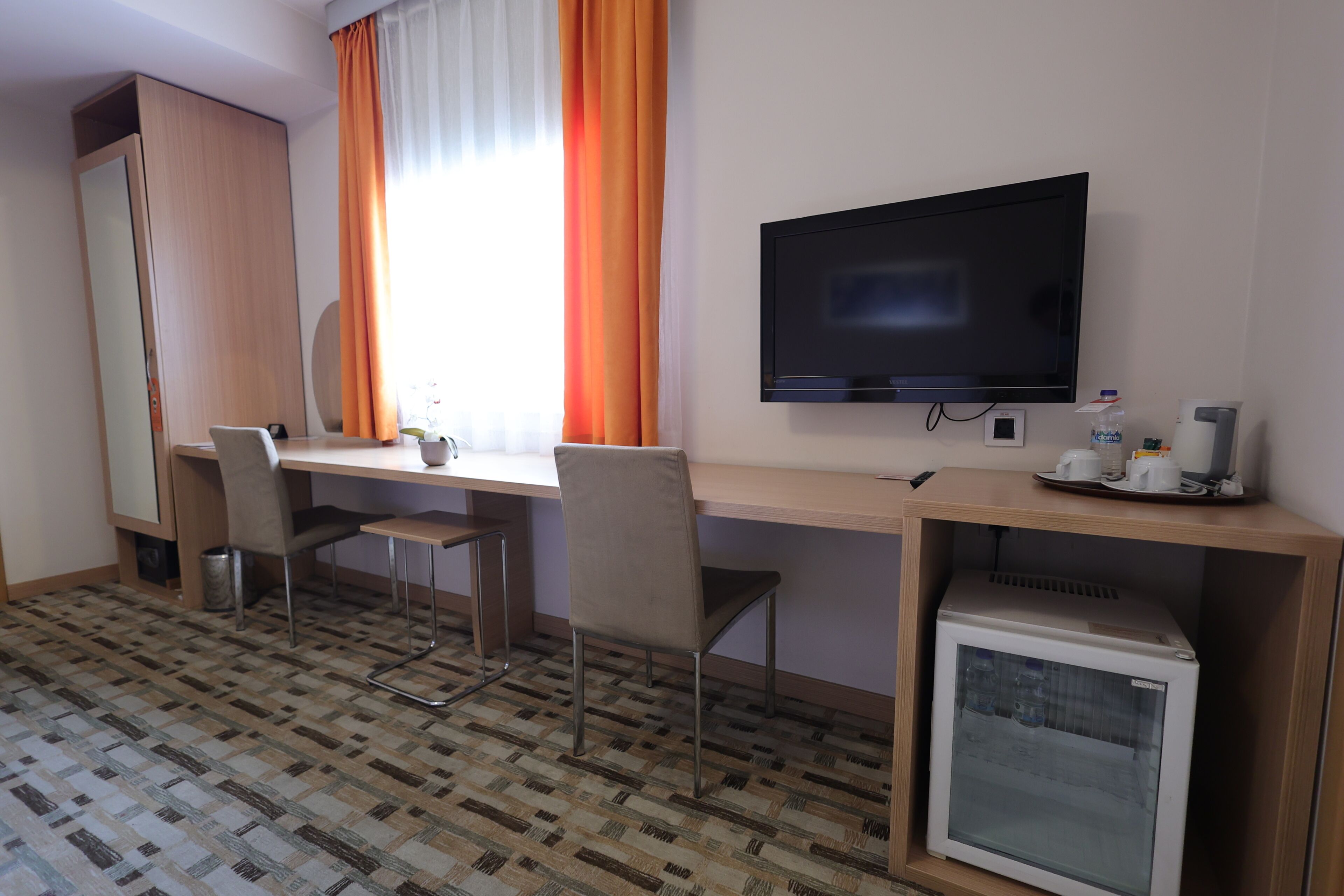A hotel room with a desk, two chairs, a television, a mini-fridge, and a wardrobe.