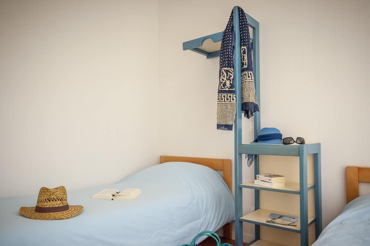 Een studentenkamer met twee eenpersoonsbedden, een blauw stapelbed, een geweven hoed, een turquoise tas en een boekenkast met boeken en een paar brillen.