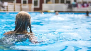 Una piscina con una persona nadando, un edificio de techo rojo y una sombrilla blanca al fondo.