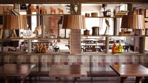 Un interior rústico con mesas y bancos de madera, estanterías con diversos objetos y una vista a otra habitación.