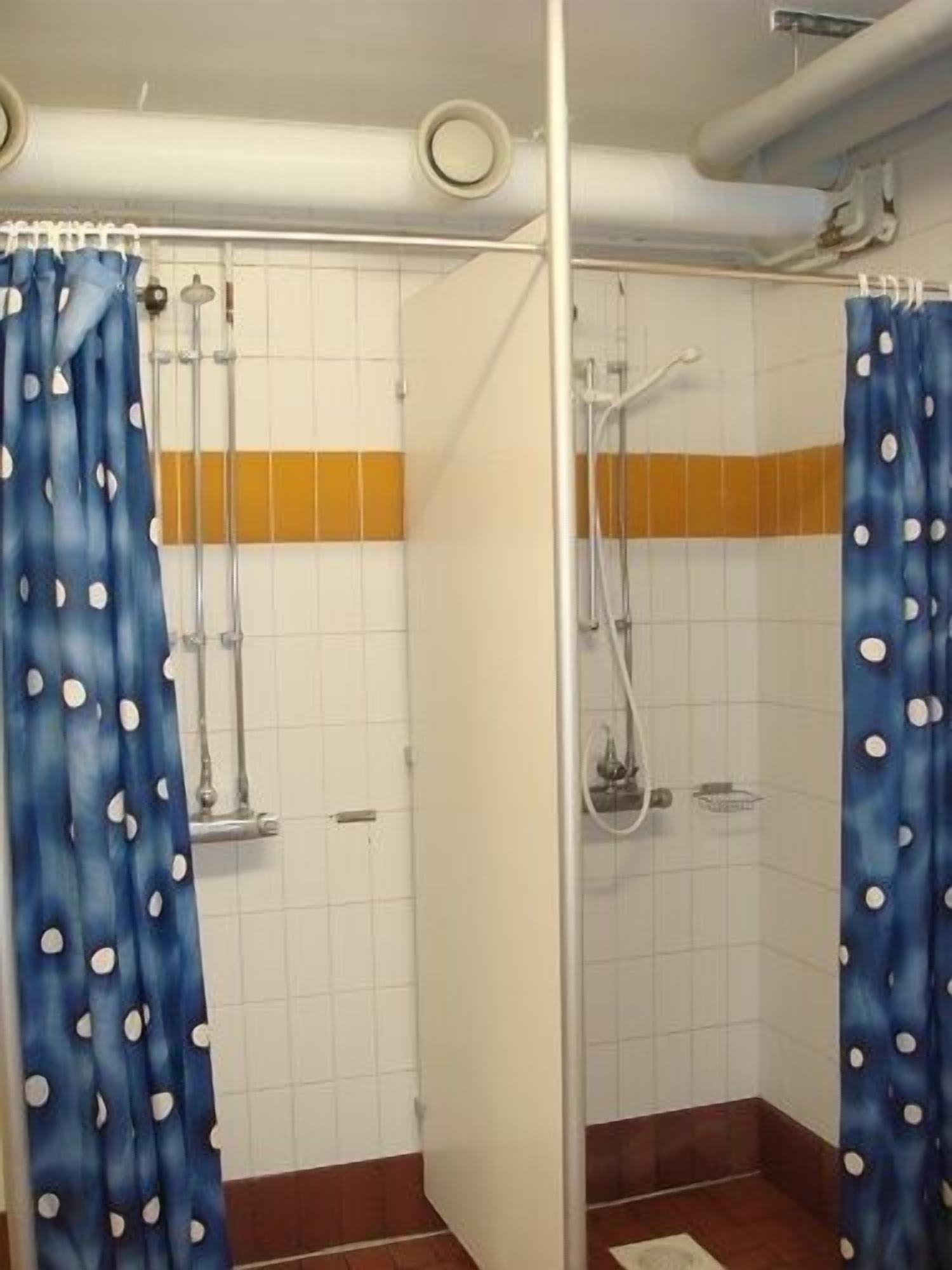 Baño con paredes de azulejos blancos, piso marrón y zona de ducha con cortina azul con lunares blancos.