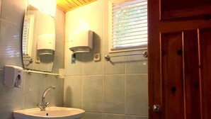 Un baño con lavamanos, espejo, dispensador de jabón en la pared y una ventana con persianas.