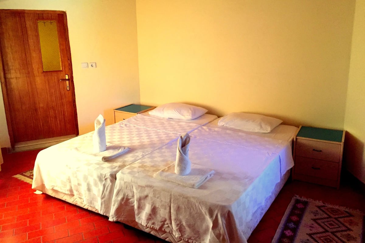 A neatly made bed with towels and a checkered bedspread, a wooden door, a small bedside table, and a wall-mounted heater.