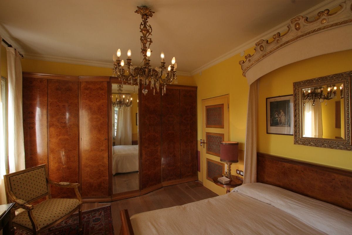 A hotel room with a large bed, a chandelier, a wardrobe, a chair, and a mirror on the wall.