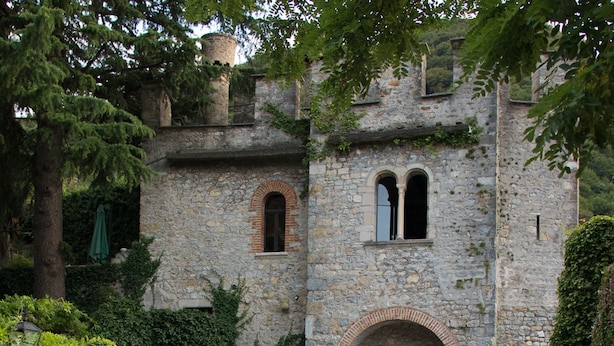 Un edificio storico in pietra con un ingresso ad arco, circondato dal verde e da un cortile in ghiaia.