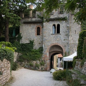 A historic stone building with an arched entrance, surrounded by greenery and a gravel courtyard.