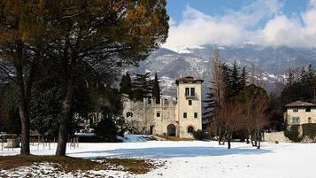 A stone building with a tower, surrounded by snow-covered ground and trees, with mountains in the background.