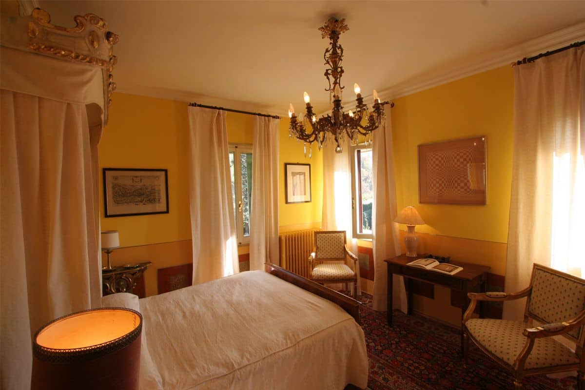 A hotel room with a large bed, a chandelier, a desk with a lamp, two chairs, and a view of the outdoors through the windows.