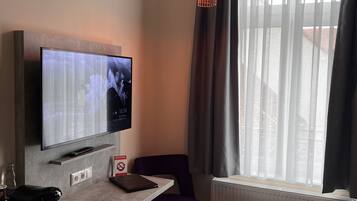 A modern hotel room with a flat-screen TV mounted on the wall, a small table with a chair, a coffee maker, and a window with sheer curtains.