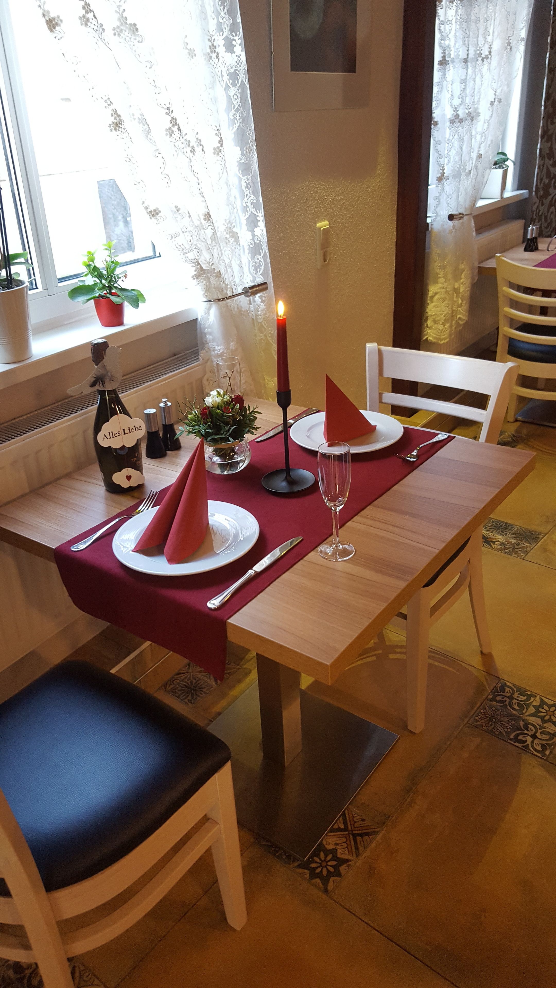 A dining table set with plates, cutlery, a candle, and a glass. There are chairs around the table, and a window with curtains in the background.