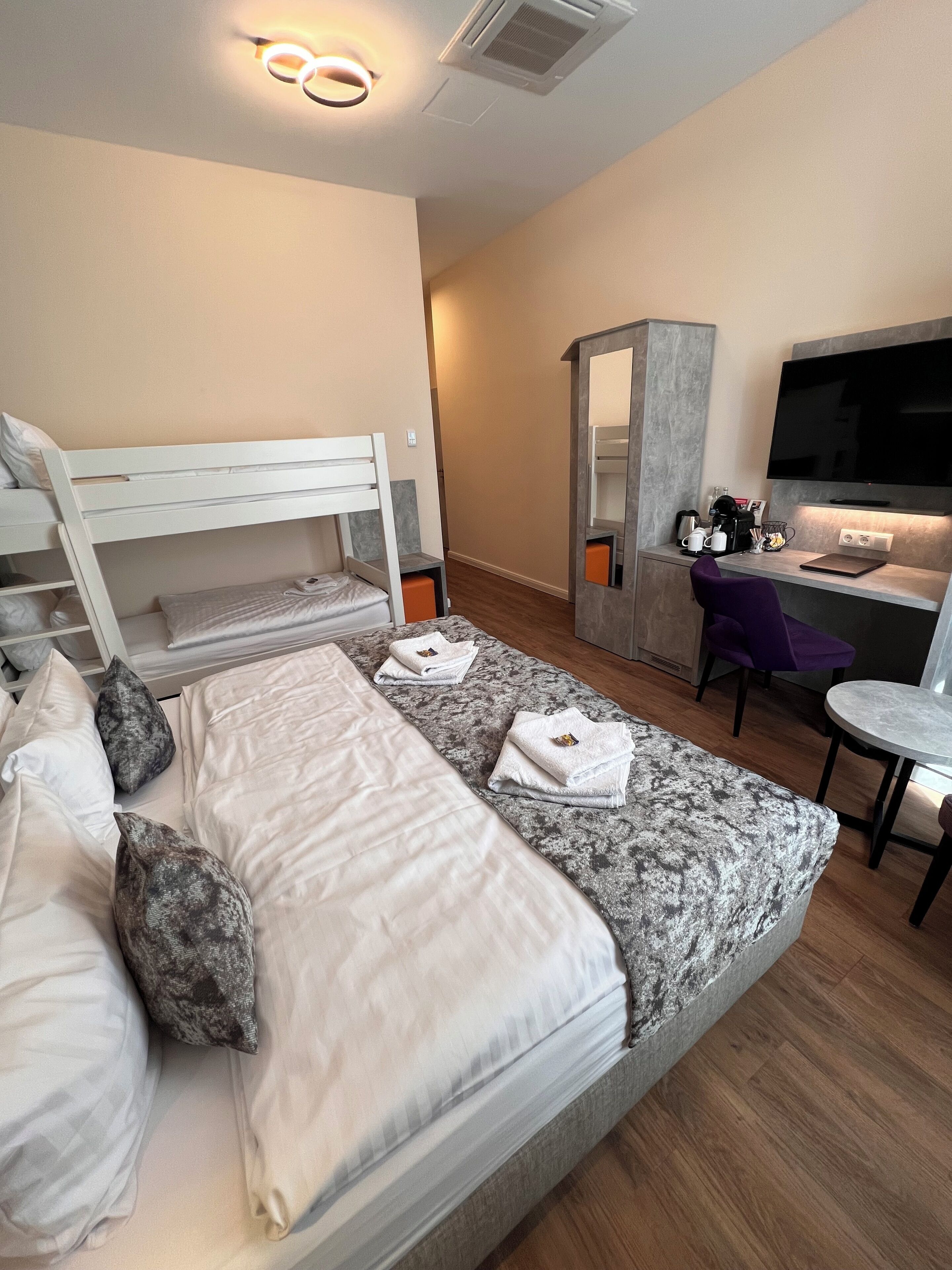 Una habitación de hotel moderna con una cama individual superpuesta, un escritorio con silla, un televisor y una pequeña zona de estar.