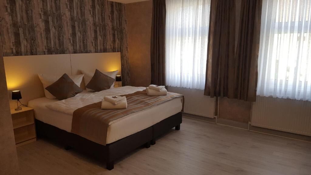 Habitación de hotel moderna con una cama grande, una mesita de noche, una lámpara y vistas al exterior a través de cortinas translúcidas.