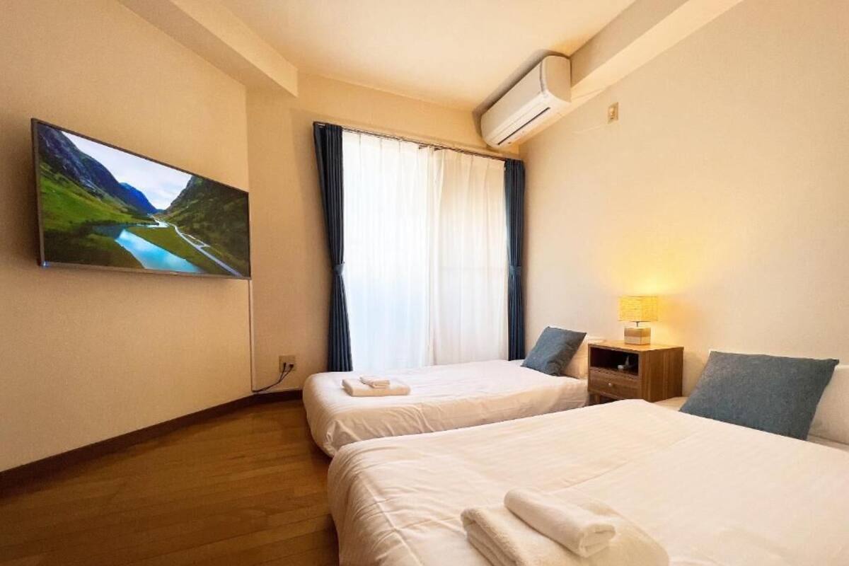A hotel room with two beds, a TV on the wall, a window with curtains, an air conditioner, and a bedside table with a lamp.