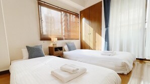 Quarto de hotel com duas camas, piso de madeira, mesa de cabeceira e janela com persianas.