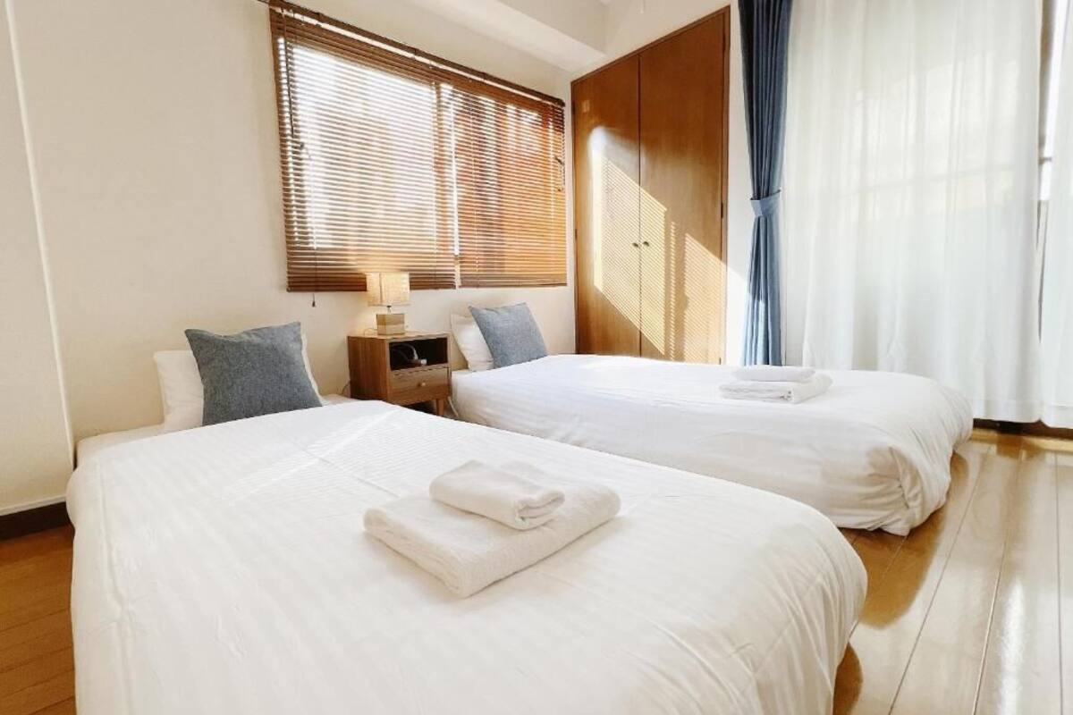 A hotel room with two beds, wooden flooring, a nightstand, and a window with blinds.