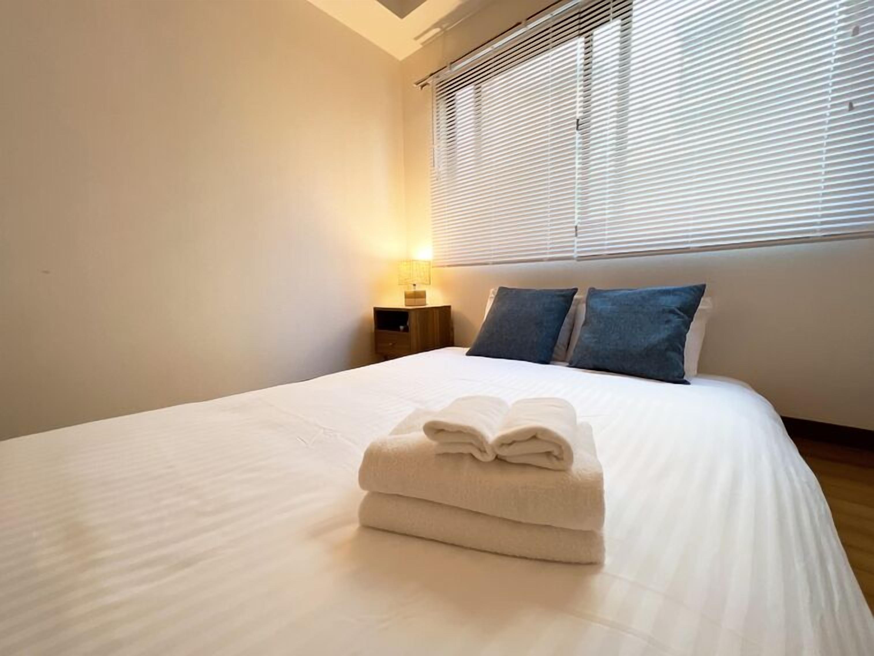A neatly made bed with folded towels, a bedside lamp, and a window with blinds.