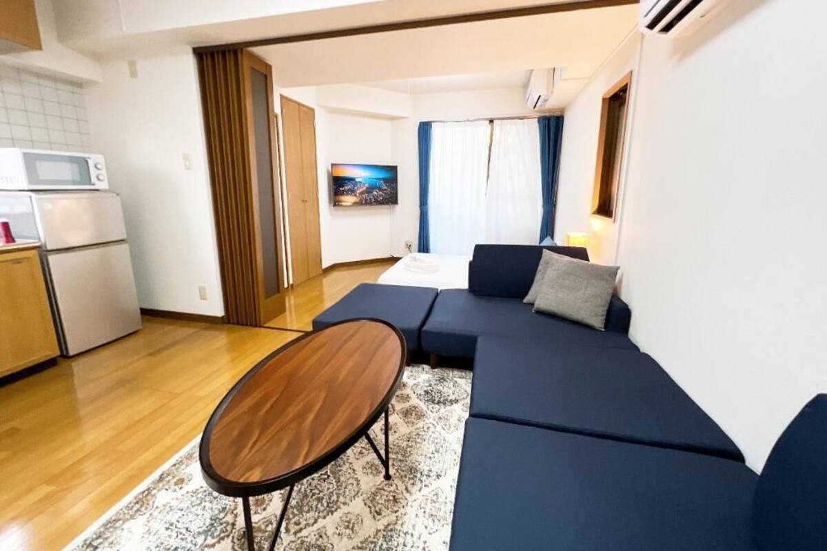 A modern living room with a dark blue sofa, a wooden coffee table, and a kitchen area with a microwave and refrigerator.