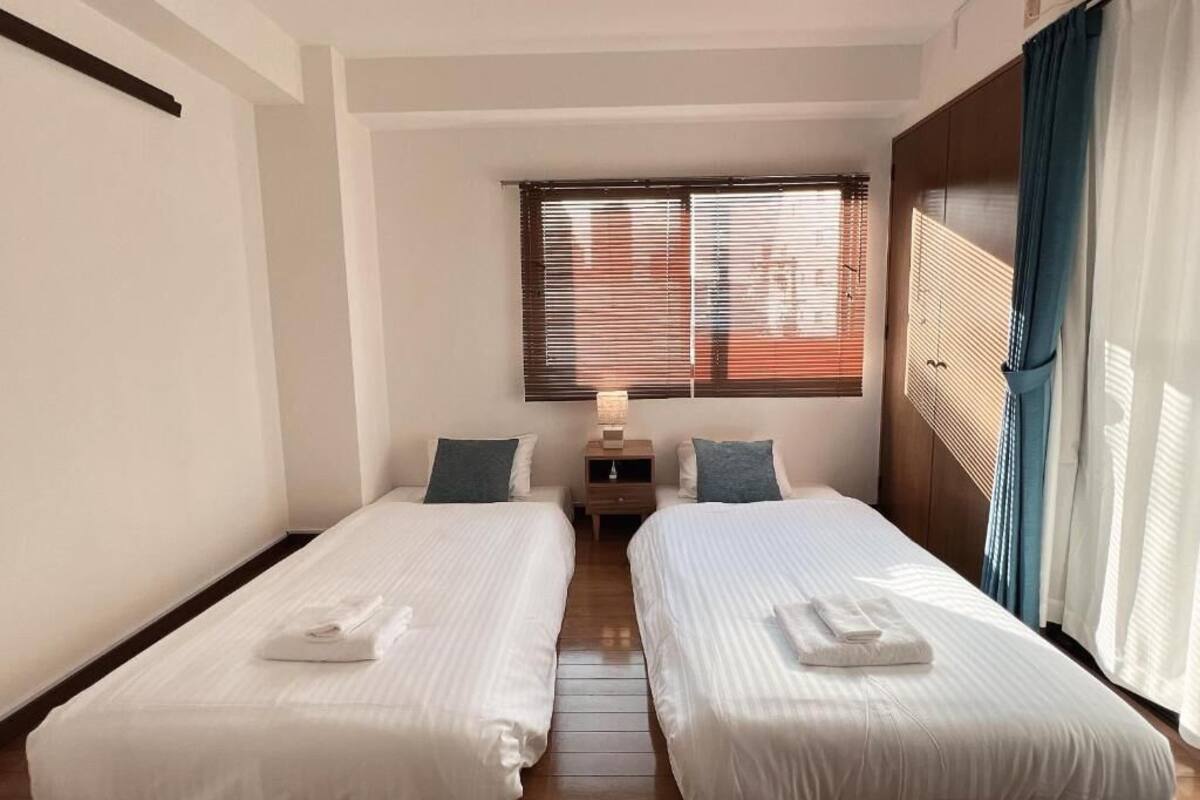 A hotel room with two single beds, a nightstand, a lamp, and a window with blinds.