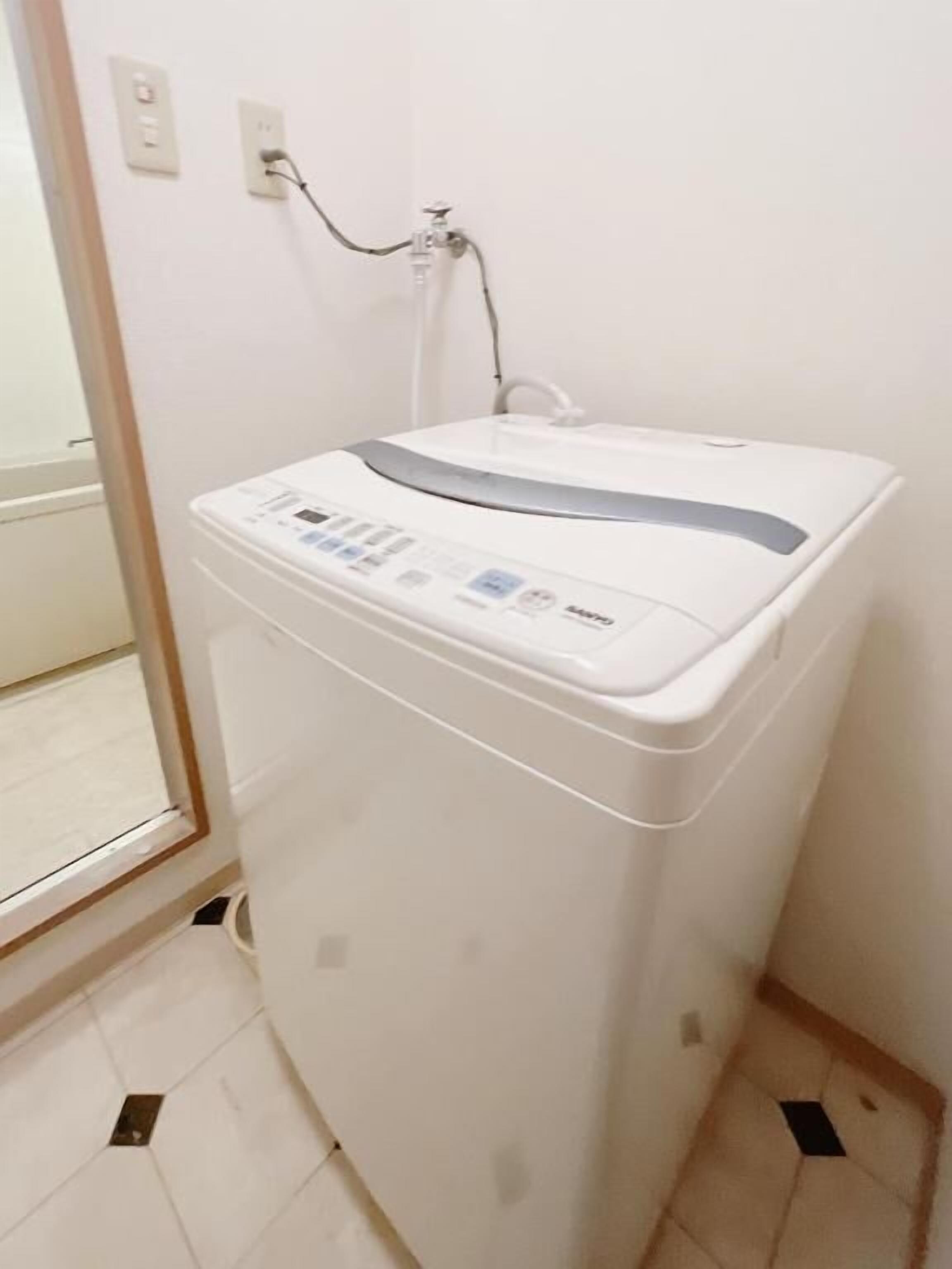 A compact, white washing machine with a control panel, located in a corner of a room with a tiled floor and a door leading outside.