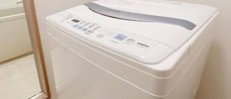 A compact, white washing machine with a control panel, located in a corner of a room with a tiled floor and a door leading outside.