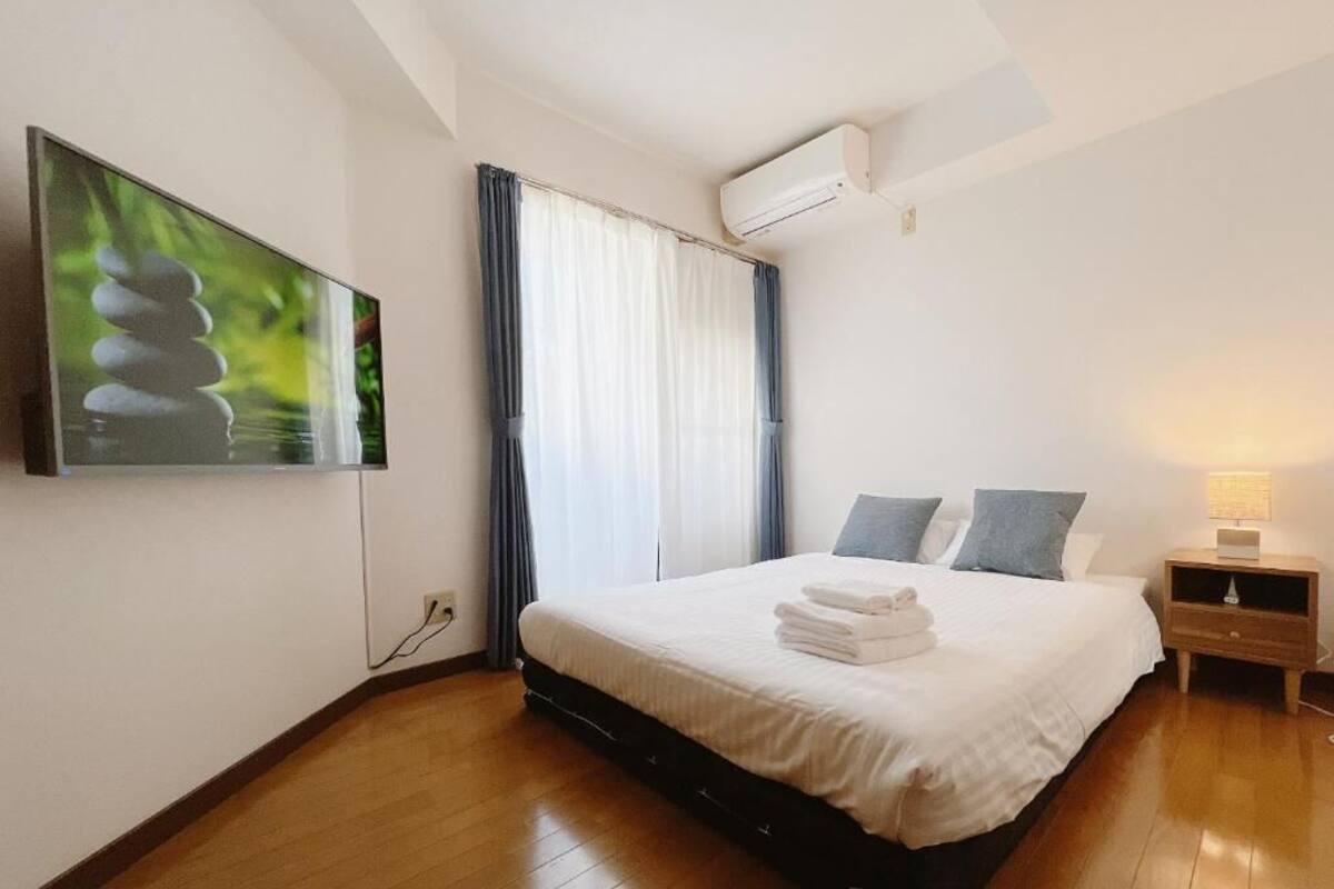 A bedroom with a bed, a TV mounted on the wall, an air conditioning unit, a bedside table with a lamp, and a window with curtains.