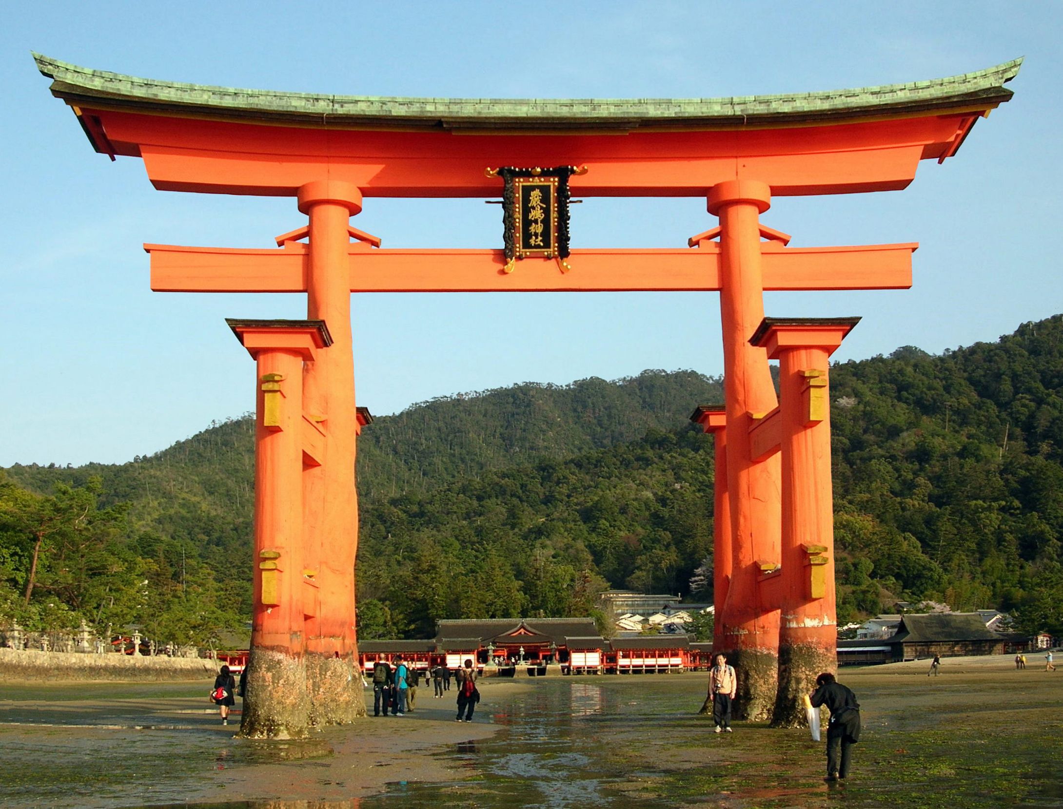 A traditional Japanese torii gate with a green roof, surrounded by a forested area and a small shrine in the background.