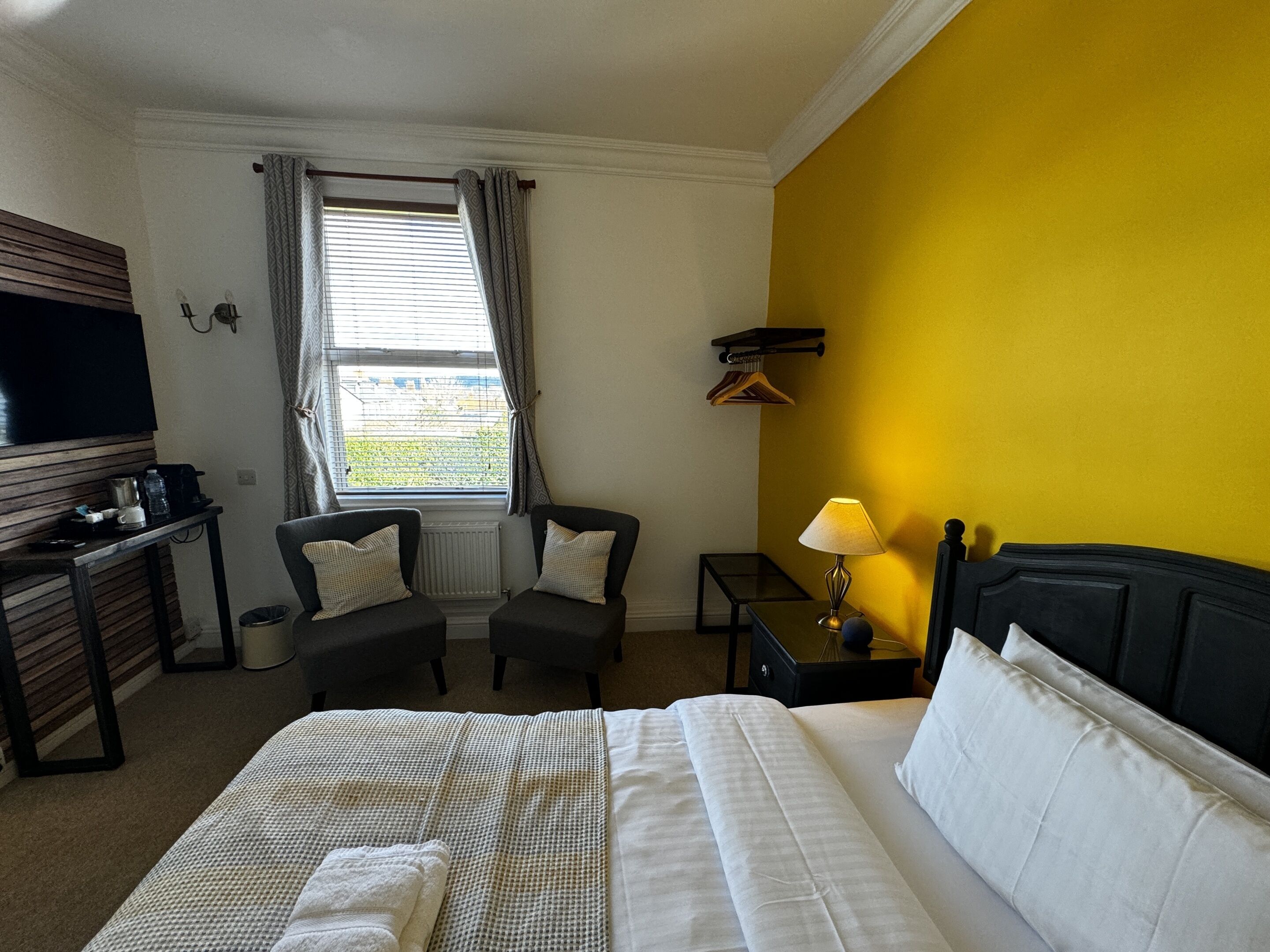 A hotel room with a bed, two armchairs, a small table with a lamp, a TV, and a window with curtains.