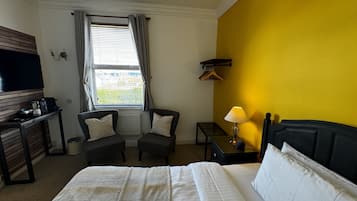 Quarto de hotel com uma cama, dois poltronas, uma mesinha de centro com abajur, televisão e janela com cortinas.